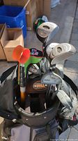 View of golf clubs standing in a black golf bag showing some club heads and club grips. Orange and black golf handle visible.