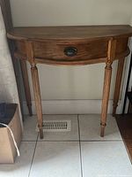 Front view of a curved semi-circular wooden hall table with a small central drawer and four slender fluted legs, placed against a white wall on tiled floor.
