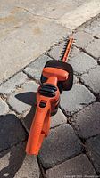 Orange and black electric hedge trimmer lying on a stone-paved surface, showing the handle, trigger, and blade in close-up.