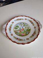 Full view of the round bone china plate with handles on both sides and detailed decoration including bird, flowers, gold trim, and red border.