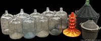 Eight large glass carboys arranged in a row, with two bottle drying racks: an orange plastic one with multiple pegs and a gray basket style. One carboy is protected by a green wire basket. Items appear used and dusty.