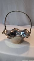 Front and side view of a silver basket showing its scalloped edges and twisted wire handle against a plain background.