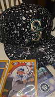 Close-up of the black Mariners cap with a white splatter design and embroidered Mariners 'S' logo, accompanied by a 2002 Ichiro Topps rookie card.