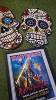 Photo showing the framed Iron Maiden Run For Your Lives World Tour 2025-26 concert poster on a green surface with two decorative Day of the Dead style skull objects nearby.