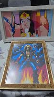 Two framed art prints shown together, one pop art print featuring blonde woman in superhero dress, one fantasy skeleton warrior print in wood frame.