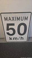 Front view of large metal speed limit sign with 'MAXIMUM 50 km/h' reflective text on white background and minor wear