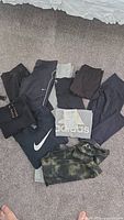 Photo showing the 10 activewear items laid out on floor, featuring black Nike pants, grey Adidas shirt, camo long-sleeve, and other black and grey workout clothes.