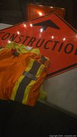 Orange diamond-shaped construction sign and three safety work shirts, two yellow with reflective stripes, and one orange.