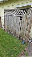 Wooden fence panel with lattice top leaning against a yellow siding wall on grass, showing overall condition and size.