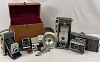 Photo of three vintage Polaroid cameras, multiple flash attachments, a small light meter and carrying case arranged on a white surface.