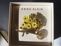 Close-up of gold-tone Anne Klein pin in the shape of a wheelbarrow filled with yellow enamel sunflowers and green leaves, edged with rhinestones, placed in original branded box.