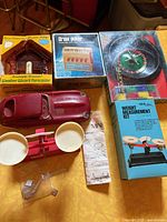 Photo of assorted vintage items including boxed Weather Wizard Forecaster, Draw Poker game, roulette wheel, weight measurement kit, red toy car, and small balance scale.