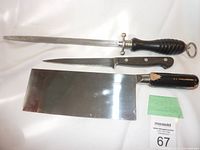 Photo showing three cutting instruments: a long sharpening steel, fillet knife with wooden handle, and a large cleaver with marked blade.