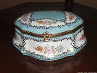Front view of hand painted Limoges porcelain trinket box with floral designs on blue and white background, gold accents, and metal trim clasp.