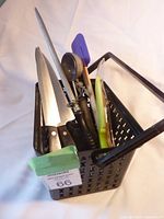 Photo of black perforated metal container holding assorted kitchen utensils including spoons, spatula, knives, peeler, and corn holder.