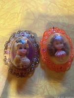Two vintage Lucky Kiddie Lockets side by side, showing the orange frame and clear frame with fogged domed covers and figurines inside.
