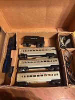 Top view of the full set in a cardboard box showing the blue locomotive, black tender car, three silver passenger cars, and transformer box with wiring.