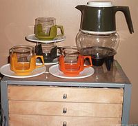 Vintage coffee/tea set displayed on a wooden chest, showing the glass pot with green plastic lid and handle, four glass cups with colorful plastic holders on white saucers, and the warming base below the pot.
