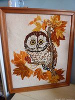 Framed needlework picture of an owl with yellow eyes sitting on a branch surrounded by orange and yellow leaves.