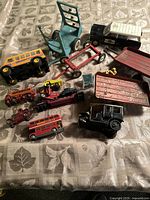 Overview of various die cast and plastic vintage toy vehicles on a patterned fabric surface, including buses, tractors, trailers, and earth movers.
