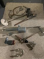 Photo showing 11 antique farm and household tools laid on carpet including clamps, hand drills, horseshoe, leather glove