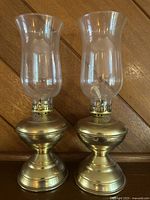 Pair of brass oil lamps with clear glass chimneys shown side by side on a wooden surface.