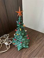 Full view of ceramic Christmas tree with colorful light bulbs and orange star topper on wooden surface with power cord visible