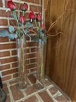 View of two clear glass floor vases standing on a brick surface near a wooden wall, each containing artificial long stem red roses, green leaves, and twisted branches.