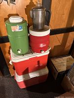 Full view showing two plastic beverage coolers, two red and white plastic ice chests, and stainless steel percolator coffee pot on top.