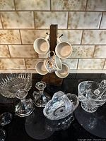Wide view of all crystal glassware and coffee mugs on a wooden mug tree on a black surface with tiled backsplash