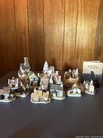 Group of 14 miniature David Winter Cottages arranged in front of wooden paneling, showcasing various architectural styles and colors in collectible painted ceramic.