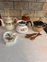 Photo showing full lot of farmhouse kitchenware items on a countertop: teapots, stoneware pitcher and jug, wooden spoons, and ceramic rooster spoon rest