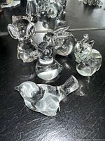 Photo showing five clear glass animal figurines including owl, rabbit, bird, snail, and elephant on a dark reflective surface.