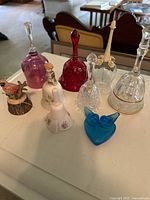 Shows all bells including pink, red, clear with flowers, tall clear ridged bell, white ceramic bell with figurine, two blue glass love birds, and two bird figurines on wooden bases.