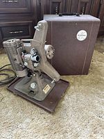 Vintage Keystone 8MM film projector side view showing lens, mechanical controls, and textured brown finish next to original carrying case.