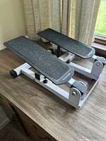 Manual stepper exerciser on wooden surface near window with white metal frame and black textured foot pedals.