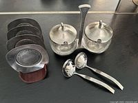 Set of two stainless steel and glass servers, two serving spoons, six stainless steel coasters, and their wooden holder arranged on black surface.