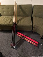 Four pool cue sticks leaning against olive green sofas, with a red case on the floor containing two cues.
