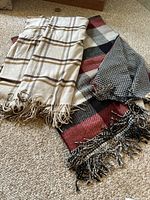 Two fringed throw blankets laid out on carpet. One is darker with multi-color plaid, the other lighter with a beige plaid pattern.