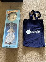Photo showing Precious Moments doll kit box and Apple tote bag side by side on carpet.