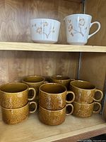 Photo showing 8 of the 10 brown Corning Cornerstone soup mugs with embossed floral patterns stacked in two rows.