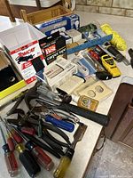 Photo showing a wide assortment of tools including screwdrivers, hammer, pliers, electrical tape, light bulbs in boxes, string, and some hardware items all loosely organized.