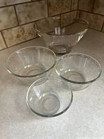 Clear view of all four glass bowls on countertop; largest clear glass bowl behind three nested Anchor Hocking bowls.