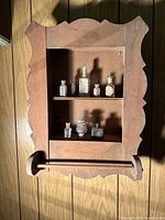 Photo of vintage wooden bathroom cabinet with scalloped edges, two shelves inside and towel bar at bottom, showing seven miniature bottles on shelves.