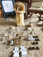 Wide view of large angel doll sitting on carpet holding a candle, surrounded by assorted smaller angel ornaments, figurines, cutouts, and a book on the floor.