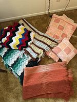 Five vintage crocheted afghan blankets in various patterns and colors with a matching pink crocheted pillow shown on carpeted floor.