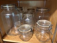 Seven clear glass vintage canning jars arranged on a wooden shelf showing varying sizes and metal clasp lids.