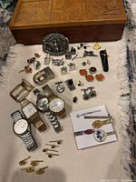 Full lot of vintage jewelry pieces laid on cloth beneath carved wooden box, showing watches, pins, cufflinks, belt buckles, and tie clips scattered loosely.