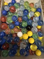 Close-up view of vintage glass marbles of assorted colors inside a white box, showing variety of internal swirl patterns and color blends.