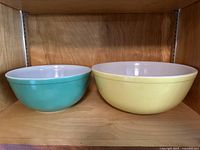 Two vintage Pyrex mixing bowls side by side on a wooden shelf - yellow #404 and green #403 bowls, showing their exterior colors and white interiors.